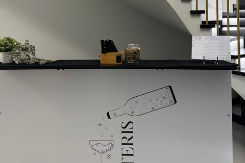 Stilingas baltas baro stalas su minimalistiniu užrašu „ETERIS“, dekoruotas grafiniu butelio ir taurė