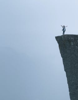 Woman standing on a tall cliff with her hands spread wide. Is it a photoshoot or will she jump?