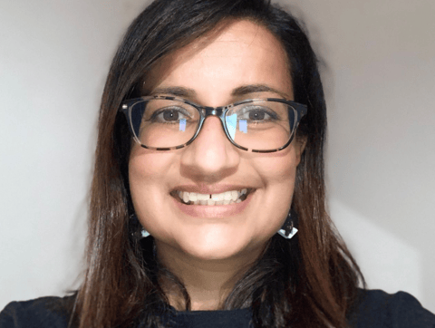 Photo of a brown lady with long dark hair, wearing glasses and a dark top, smiling against a light b
