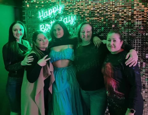Friends in front of the rose gold shimmer wall with green βHappy Birthdayβ neon for Mae's 33rd birthday in Redlands.