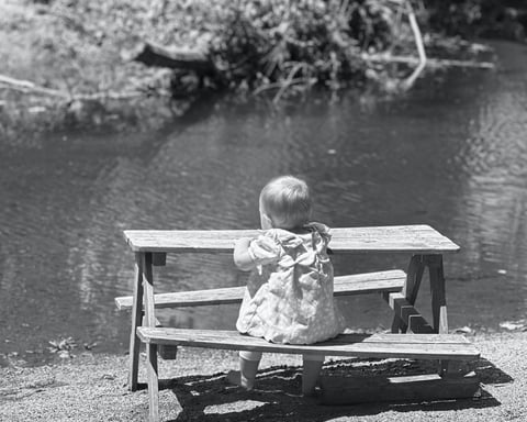 Black and white photo of a baby girl sitting at a miniture picnic table, gazing at a lake with trees in the distance.