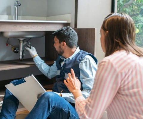 plumber pointing under a sink and explaining things to a woman