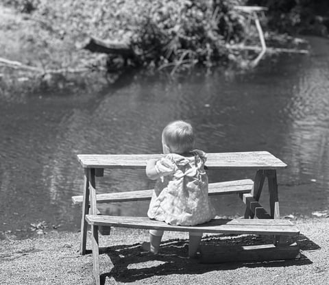 Black and white photo of a baby girl sitting at a miniture picnic table, gazing at a lake with trees in the distance. 