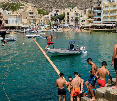 Men and boys in swimming shorts gather on the edge of Xlendi Bay by a greasy pole that juts out over the water.