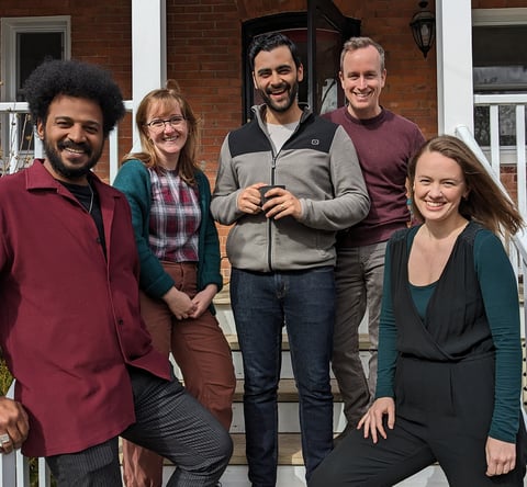 A group of five diverse artists pose on the steps of a brick house, smiling at the camera