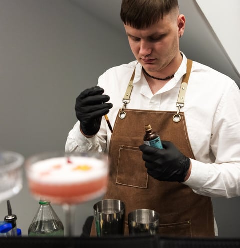 Barmenas, dėvintis baltus marškinius ir rudos spalvos prijuostę, ruošia kokteilį profesionalioje bar