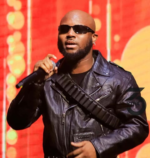King Promise in a black vest holding a microphone in dark spectacles.
