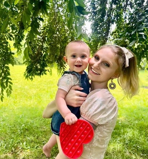 Mother holding baby close under a tree, both smiling at the camera, baby holding heart-shaped toy.