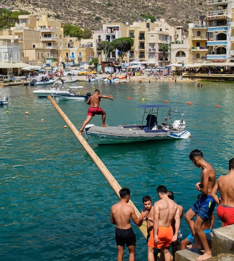 Men and boys in swimming shorts gather on the edge of Xlendi Bay by a greasy pole that juts out over the water.