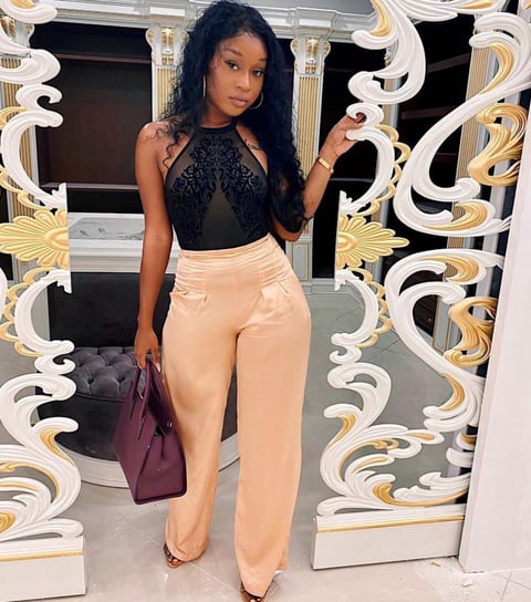 Efia Odo poses in a black net top, in a cream trousers and a mauve handbag.