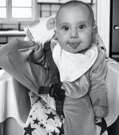 Black and white photo of smiling baby in high chair with tongue out, showing natural baby joy, innocence, and purity.