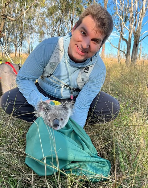 Ted Pavlic in hiking clothes holding a bag with a small koala head poking out of the bag. Eucalyptus trees in background.