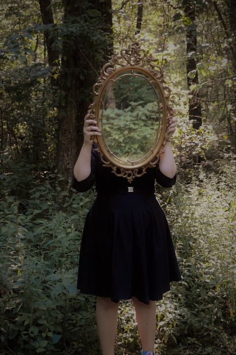 Woman holding mirror in forest, reflecting the sacred field of relationship and unseen inner truths