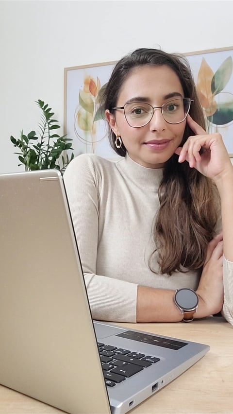 Mulher de óculos, suéter bege sentanda em uma mesa em frente a um notebook olhando para frente 