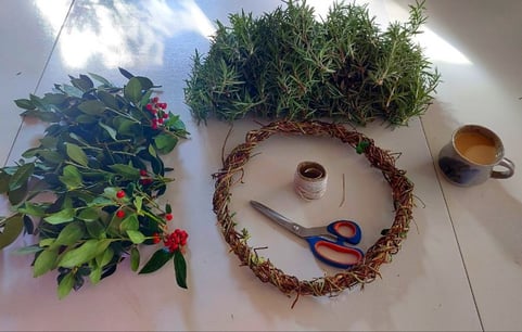rosemary and holly berry bush cuttings beside a honeysuckle wreath base with scissors and twine on a