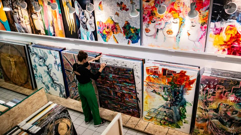 A female curator sorts through large canvas in an art gallery filled with stacks of modern paintings