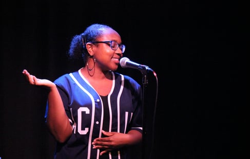 a Black woman in a navy baseball jersey and glasses speaking into a microphone