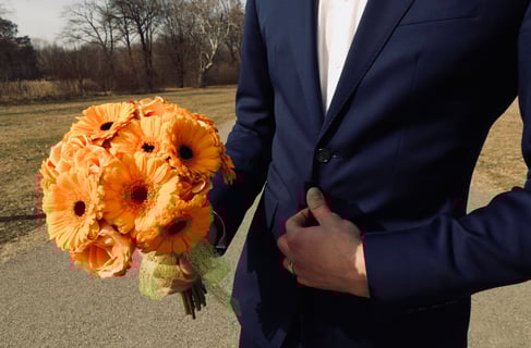 Bărbat în costum bleumarin ținând un buchet cu gerbera portocalii.