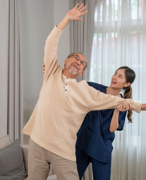 An elderly man and a younger female physical therapist in a living room doing shoulder exercises.
