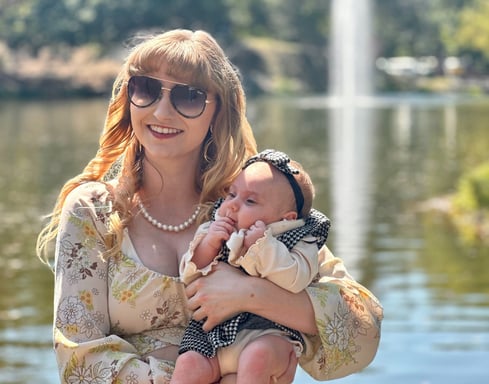 A mother cradling her newborn daughter in front of a lake, with a fountain softly spraying in the background.