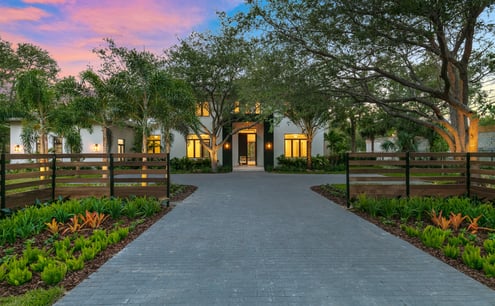 a brick driveway leading up to a well lit luxury home at sunset time with a vibrantly lit sky