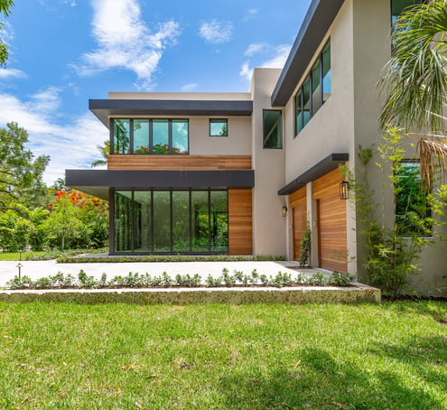 front yard of a luxury square looking home with a driveway, two garages and a forest. 