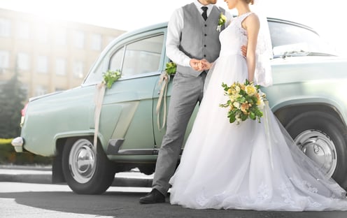 Image of a couple in wedding dress and formal suit standing beside an automobile.