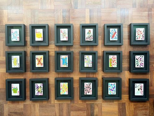vista de cima das 18 cartas de baralho personalizadas e colecionáveis da série, emolduradas em preto sobre chão de taco.