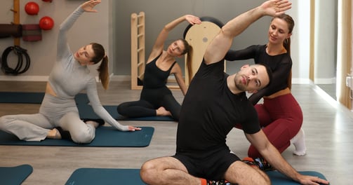 A group Pilates class performing a Pilates mermaid position on mats, with an instructor assisting.