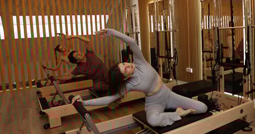 Três pessoas a fazer um alongamento lateral em reformers num estúdio de Pilates com espelhos 