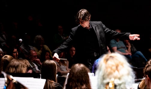 Conductor in black, with arms spread, amid heads of orchestra-members.