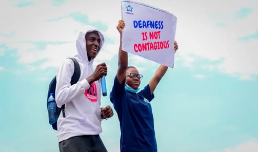 A man in a white hoodie, standing next to a woman who is holding up a banner that says "Deafness is not contagious"