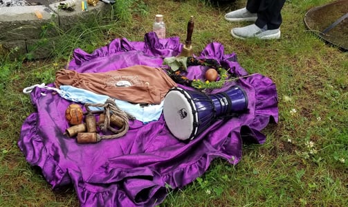 Outdoor ceremonial setup on a purple cloth with a drum, bells, moon water, zodiac cloth, and ritual
