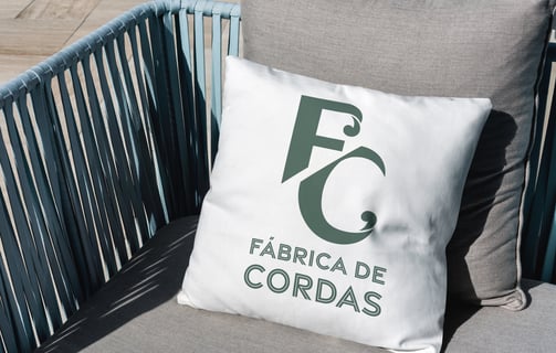 Logotipo da FC Fábrica de Cordas estampado em travesseiro sobre cadeira, destacando a matéria-prima da empresa.