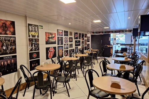 Área da cafeteria com mesas cadeiras e pôsteres de rock no Studio Brothers em Porto Alegre