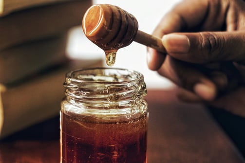 Hand holding a honey dipper dripping golden honey into a glass jar, creating a warm scene