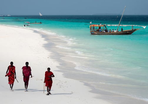 three people walking on a beach with boats in the background