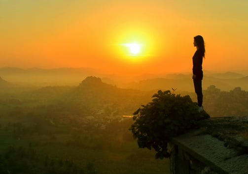 Silhouette of a woman standing calmly on a ledge overlooking a sunrise-lit valley, symbolizing peace