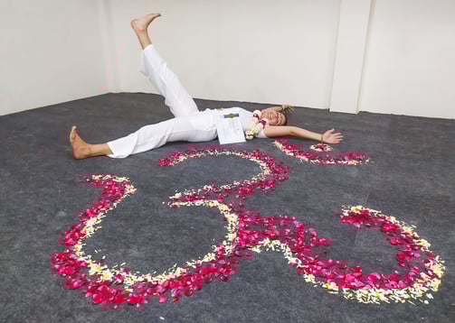 Eva lying on the ground next to a flower mandala, smiling happily while holding her Yoga Teacher Training diploma in Rishikes