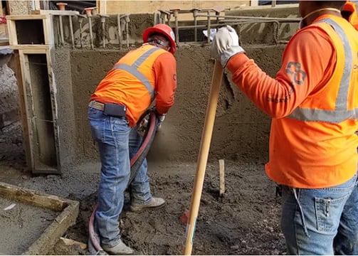 Concrete workers wearing orange safety vests and hard hats, one holds a hose, the other a shovel.