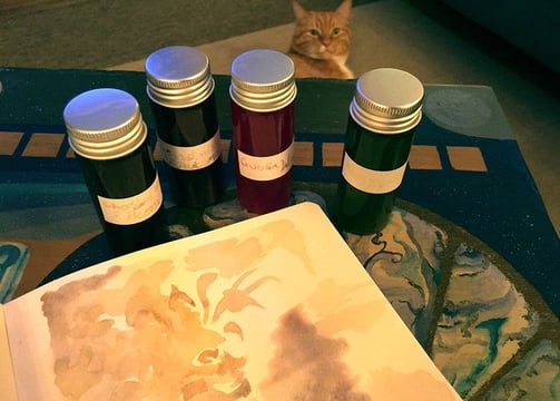 An open sketchbook with a fantasy watercolour illustration on a table surrounded by inks and a cat.