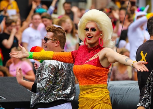 A drag queen in a rainbow dress with large blonde wig holds her arms out in greeting