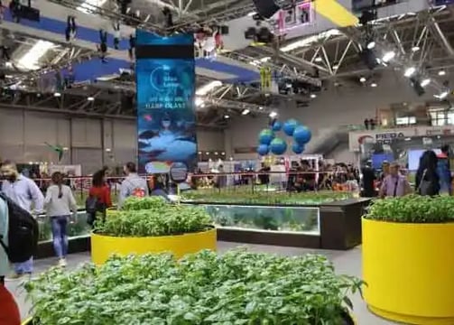 An indoor exhibition or trade show filled with people walking around various displays. Large planter