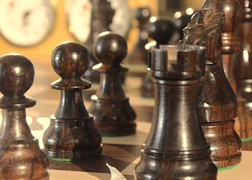 Closeup view of a chess board with black pieces and a chess clock in the background.