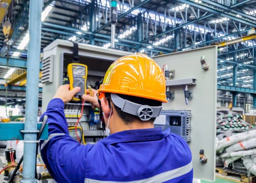 Industrial electrician wearing hard hat and gloves, servicing a large electrical panel in a warehous