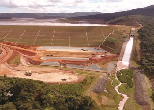 Vista aérea de uma barragem com vertedouro em funcionamento, cercada por áreas verdes e montanhas ao fundo