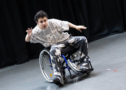 A mixed race manual wheelchair user, leaning to the side, arms spread out, and one foot in front