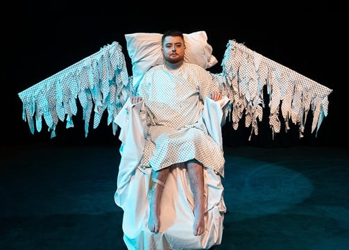 An electric wheelchair user in a hospital gown, with swooping wings made of the same fabric