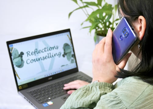 lady using mobile phone sat in front of laptop with reflections counselling on screen