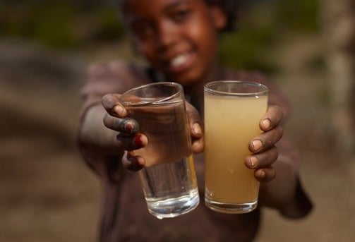 A smiling young girl holding two bottles, showing the difference between dirty and purified drinking